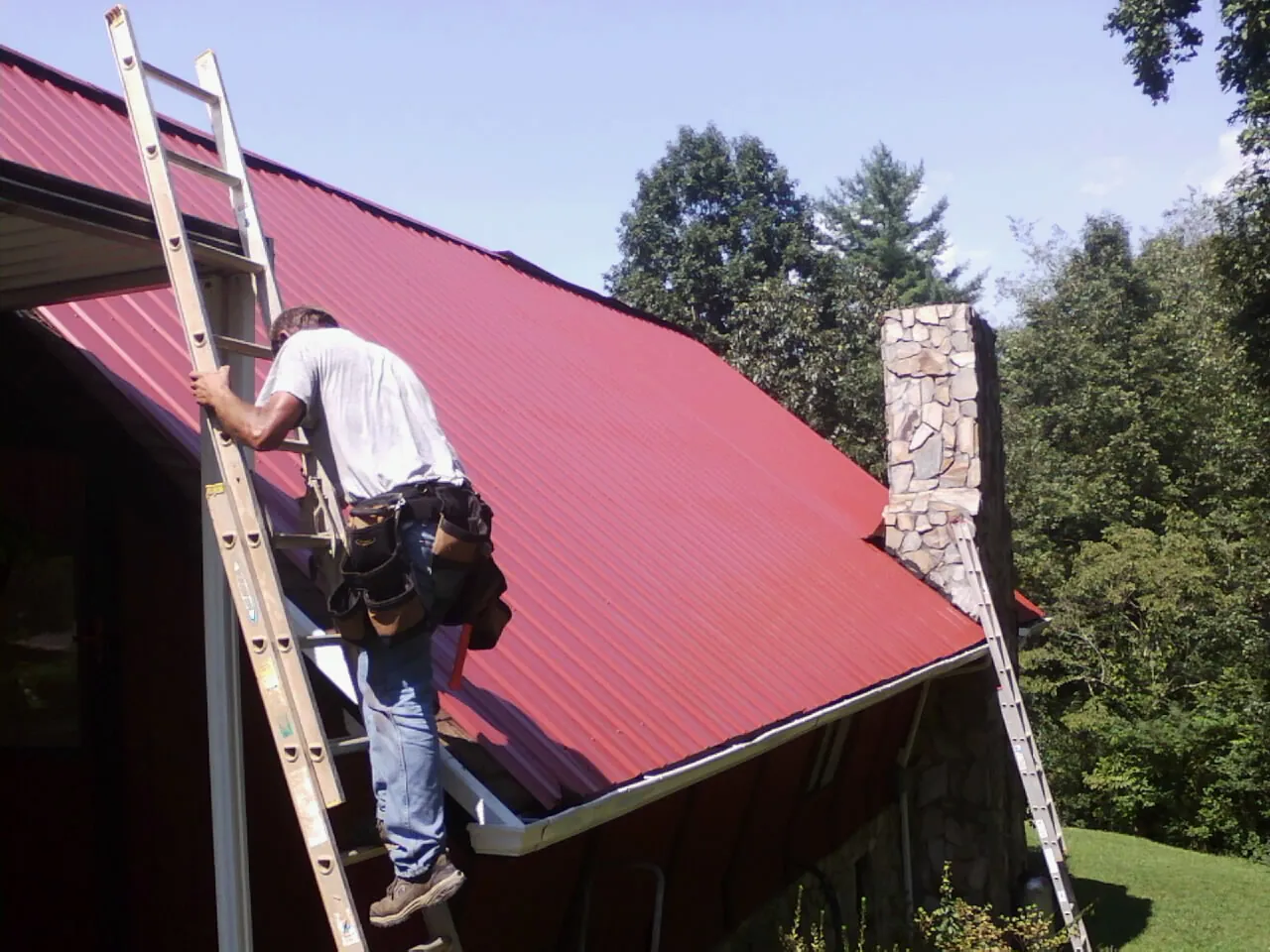 Roofer on a ladder beside a red metal roof and stone chimney — Soffit & Fascia Repair by Roofing Brothers in Cape May Court House